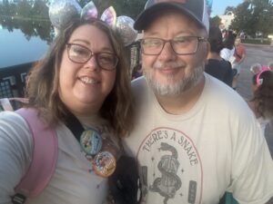 A woman wearing Minnie Mouse ears and a man wearing a hat smiling wearing their park shirts.