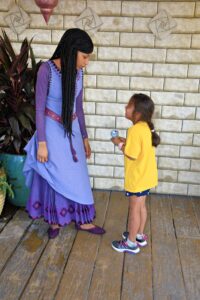 An excited girl holding a stuffed lizard looking up with joy on her face at Princess Asha in her purple dress.