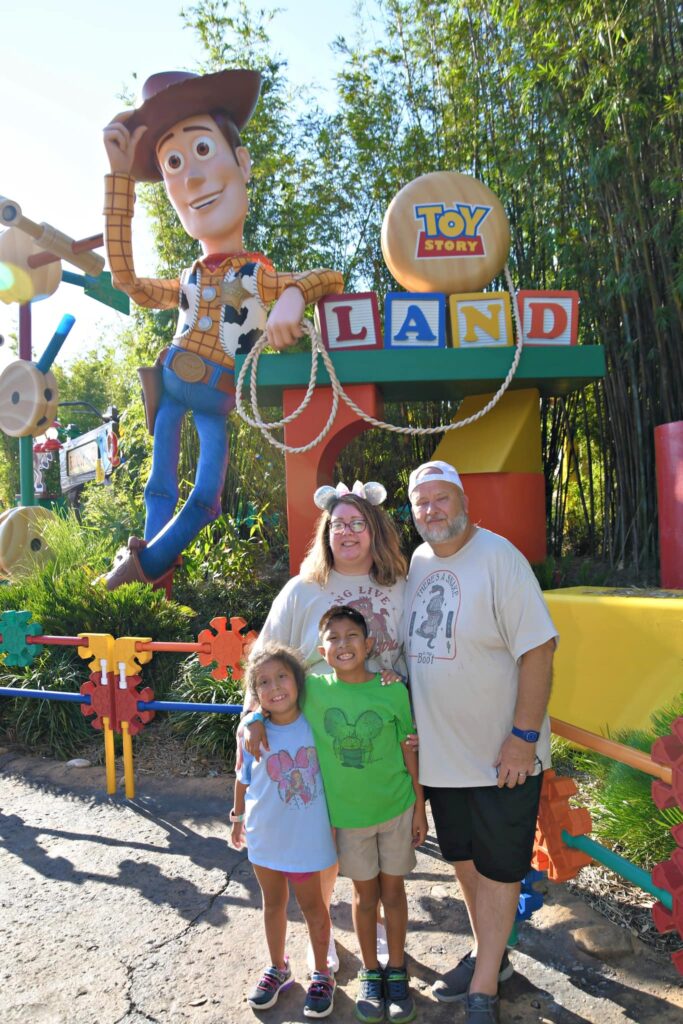 A mom wearing Disney ears and a park t-shirt, a dad wearing a backwards hat and a park t-shirt, a little girl wearing a park t-shirt, and a little boy wearing a park t-shirt smiling in front of a giant statue of Woody next to wood blocks that say Toy Story Land.