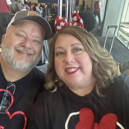 A smiling couple at an airport, the woman wearing Minnie Mouse ears with red and white polka dots and a black shirt with a red glittery heart, while the man wears a cap and a black shirt with a Mickey Mouse design. They appear excited and ready for a Disney trip, with luggage and other travelers visible in the background.